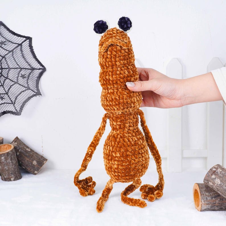 May include: A handmade, brown plush toy shaped like a quirky creature. It features a long, segmented body, small limbs, and two dark purple pom-poms. The toy is made from a soft, textured material, perfect for Halloween.