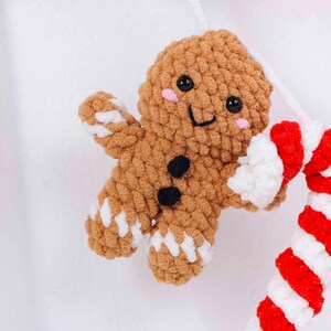 May include: A crocheted gingerbread man ornament with a smiling face, black button details, and pink cheeks. The gingerbread man is brown with white accents on the arms and legs. A red and white striped candy cane is also visible.