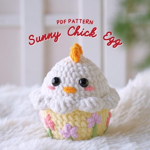 Könnte beinhalten: Ein gehäkeltes Amigurumi-Spielzeug in Form eines Küken-Eis mit gelbem Schnabel, weißem Körper und gelben und rosa Blumenakzenten. Das Spielzeug sitzt auf einer weißen, flauschigen Oberfläche. Der Text "PDF PATTERN Sunny Chick Egg" befindet sich über dem Spielzeug.