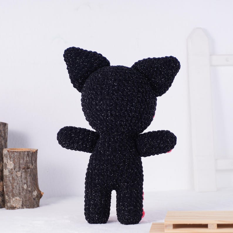 May include: A handmade black plush cat toy with large ears and outstretched arms. The cat has pink accents on its paws. The toy is made of a textured, sparkly material and stands upright.