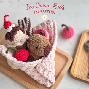 May include: A crocheted ice cream roll pattern with a pink crepe, brown ice cream, a chocolate bar, and a pink cherry. The pattern is on a wooden surface with the text "Ice Cream Rolls PDF PATTERN".