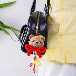 May include: A brown crocheted teddy bear keychain with a red bow, hanging from a black purse. The keychain also features a yellow bell and candy cane accents, adding a festive touch to the accessory.
