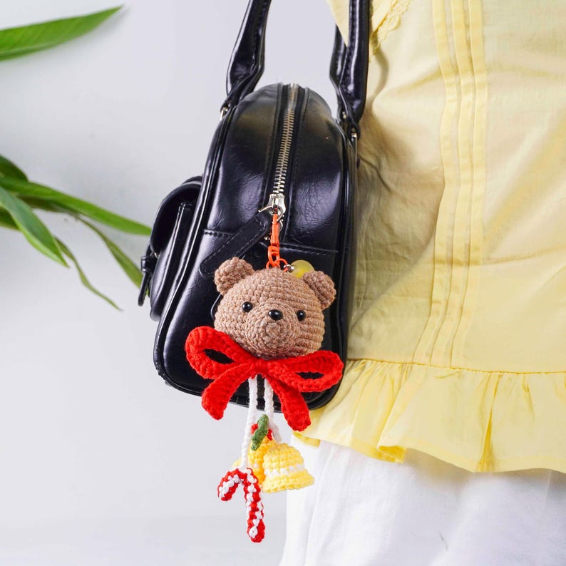 May include: A brown crocheted teddy bear keychain with a red bow, hanging from a black purse. The keychain also features a yellow bell and candy cane accents, adding a festive touch to the accessory.