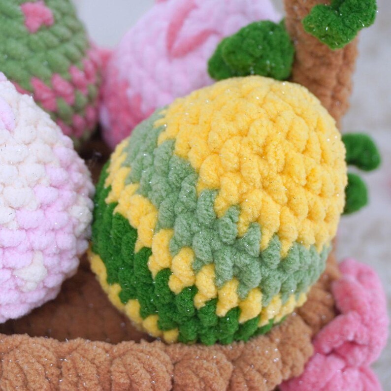 May include: A close-up of a crocheted Easter basket filled with colorful crocheted eggs. The eggs are green, yellow, pink, and white. The basket is made of brown yarn.