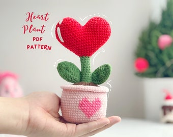 May include: A crocheted heart-shaped plant in a pink pot with a heart design. The plant is green and the heart is red. The text "Heart Plant PDF Pattern" is written above the plant.