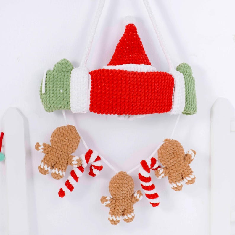 May include: A handmade Christmas decoration featuring a red and white Santa hat and a red and white candy cane design. Below, a garland of gingerbread men and candy canes hangs from the main decoration. The decoration is made of yarn.