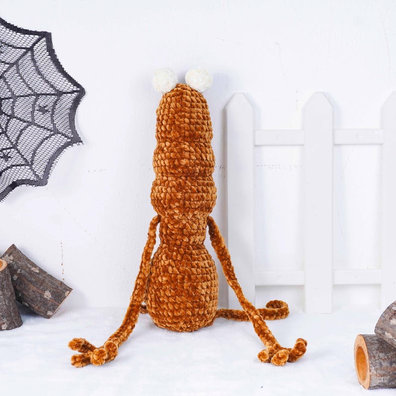 May include: A handmade, brown plush creature with a segmented body, long arms, and feet. It has two white pom-pom antennae. The plush toy is set against a white background with a decorative spiderweb and a white picket fence.