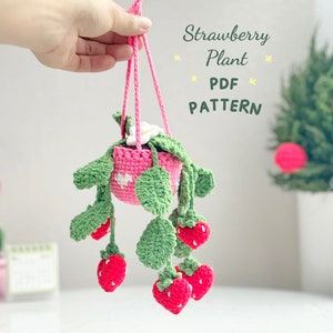 May include: A handmade crochet strawberry plant in a pink hanging basket. The plant features green leaves, white flowers, and red strawberries. The text "Strawberry Plant PDF PATTERN" is visible.