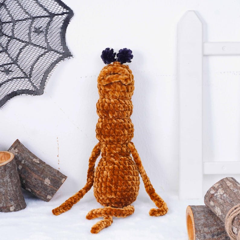 May include: A handmade, plush, brown caterpillar-like creature with two dark purple pom-poms on its head. The soft toy has long, thin arms and legs. The background includes a black spiderweb decoration and wooden logs.
