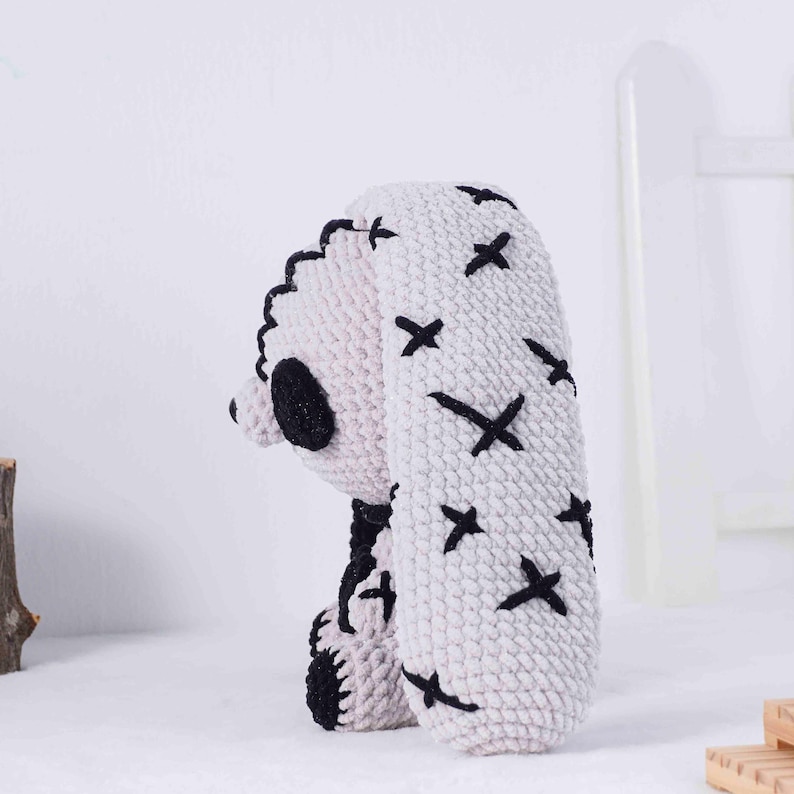May include: A handmade, crocheted bunny in a light gray color with black accents. The bunny features black cross-stitch details on the ears and a black nose and eye. The ears are long and floppy, and the overall design is soft and whimsical.