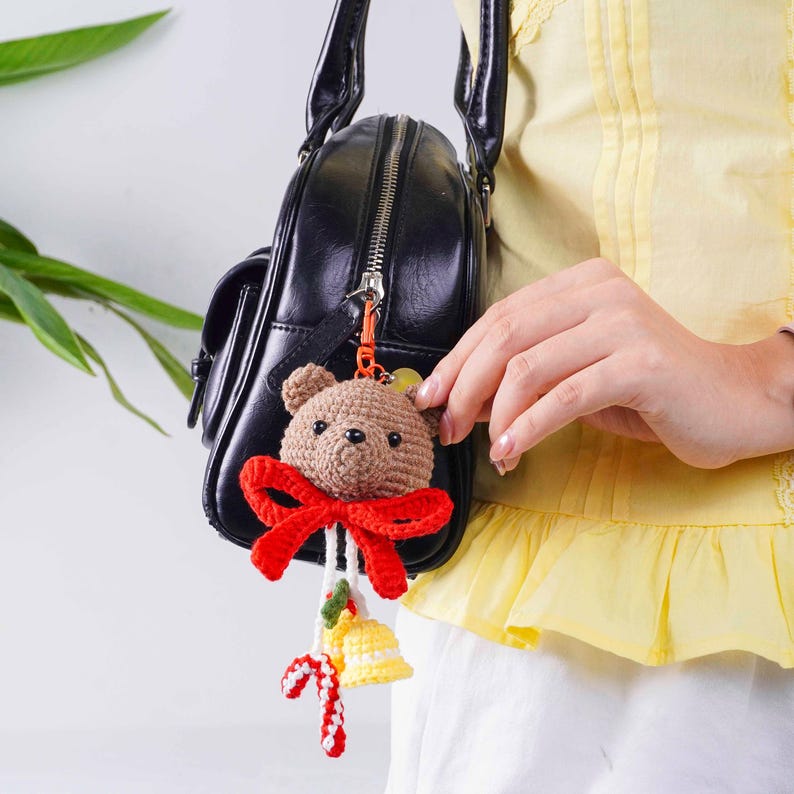 May include: A brown crocheted teddy bear keychain with a red bow, candy cane, and yellow bell charm. The bear is attached to a black purse with a silver zipper. The keychain is held by a person wearing a yellow top.