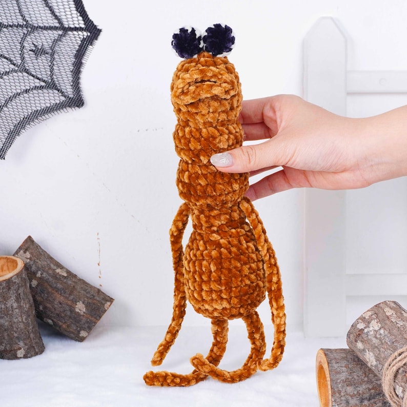 May include: A handmade brown plush toy with a segmented body and long, thin limbs. The toy has two dark blue pom-poms on top. The plush toy is held by a hand in front of a white background with Halloween decorations.