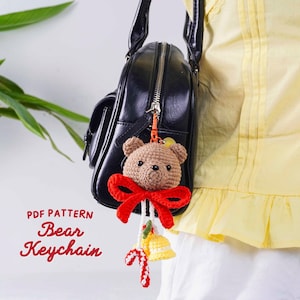 May include: A crocheted bear keychain with a red bow, hanging from a black purse. The bear is light brown with a red bow. The keychain also features a yellow bell and candy cane. Text on the image reads "PDF PATTERN Bear Keychain."