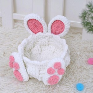 May include: A white crocheted bunny-shaped basket with pink ears and paw prints. The basket is empty and has a soft, fluffy texture.