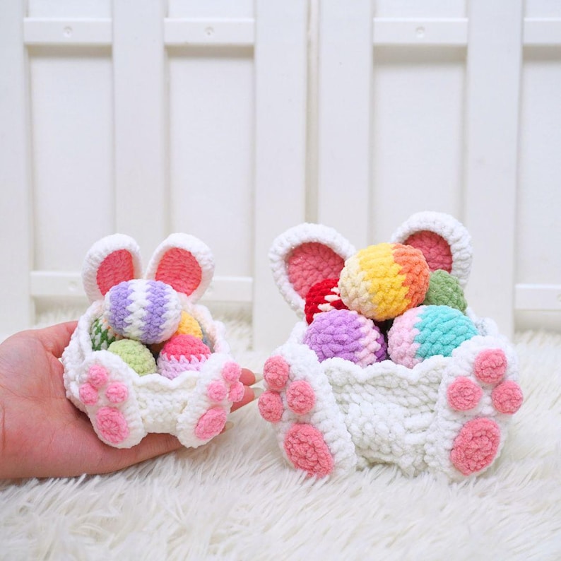 May include: Two white crocheted bunny baskets with pink ears and paws, filled with colorful crocheted Easter eggs.