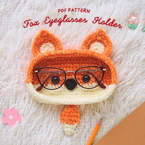 May include: A crocheted fox-shaped glasses holder with a white and orange colour scheme. The holder has a small, black nose and brown glasses.