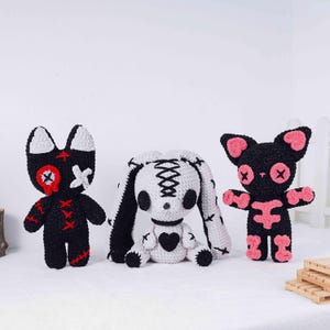 May include: Three handmade crochet dolls in a gothic style. The dolls include a black cat with pink accents, a white bunny with black details, and a black cat with red accents. Each doll has stitched details and a unique design.