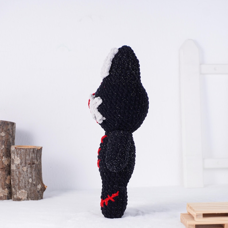 May include: A handmade black and white crochet doll with red accents. The doll has a unique design with a white face and red stitching details. The doll is standing on a white surface.