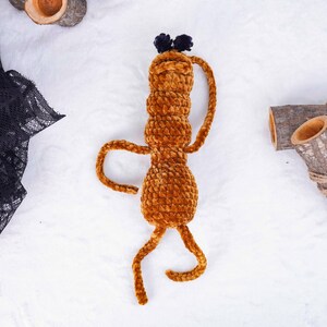 May include: A handmade, plush, brown creature with a cylindrical body, curved arms, and legs. It has a small head with two dark blue pom-poms. The toy is made of a soft, textured material, and is designed to resemble a whimsical character.