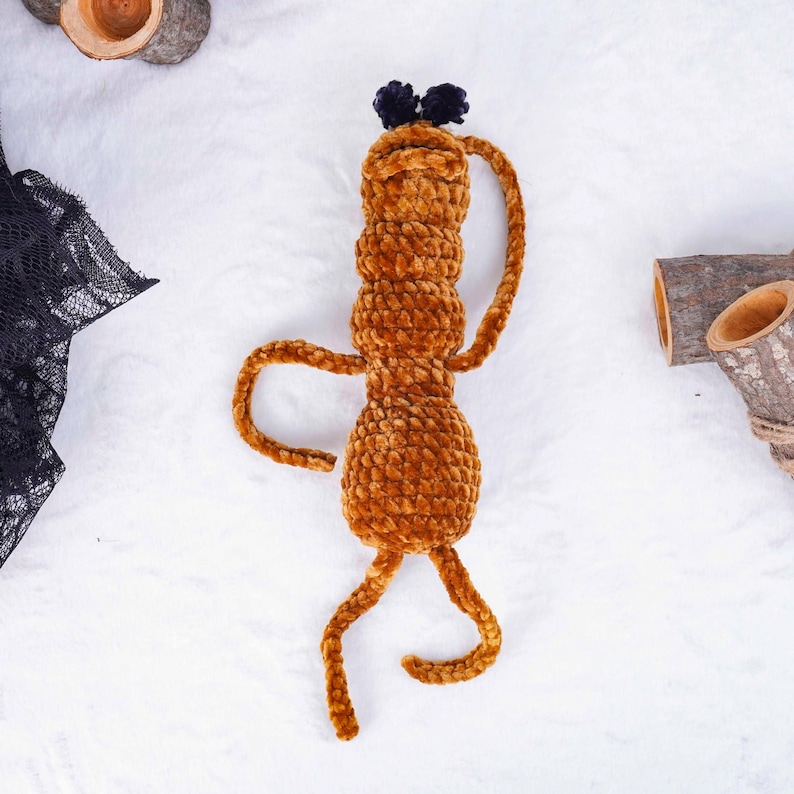 May include: A handmade, plush, brown creature with a cylindrical body, curved arms, and legs. It has a small head with two dark blue pom-poms. The toy is made of a soft, textured material, and is designed to resemble a whimsical character.