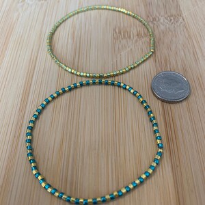 May include: Two beaded bracelets. One is gold and the other is teal and gold. The bracelets are made of small, square beads strung together to form a flexible band. A coin is next to the bracelets.