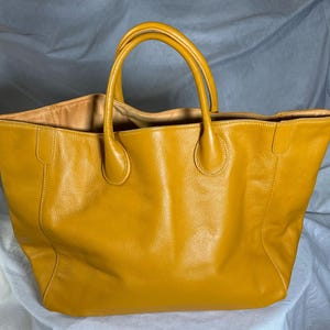 May include: A large, mustard-yellow leather tote bag with two rounded handles. The bag has a spacious interior and a textured exterior. The handles are attached with rounded, reinforced bases. The bag is designed for carrying items.