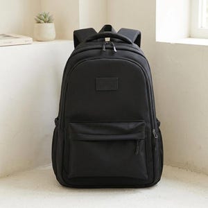 Könnte beinhalten: Ein einfarbiger schwarzer Rucksack mit einem großen Hauptfach mit Doppelreißverschluss, einer kleineren Fronttasche mit Reißverschluss und einem rechteckigen Aufnäher in der Mitte des oberen Vorderteils. Mit gepolsterten Trägern und Tragegriff.