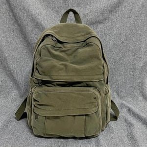 May include: An olive green canvas backpack featuring a large main compartment, a smaller front zipped pocket, and two side pockets. The bag includes a top carry handle, adjustable shoulder straps, and dark metal zip pulls for secure closure.