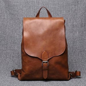 以下が含まれることがあります： 中央に縦のストラップと銀色の金属製バックルが付いた大きな折り返しフラップが特徴のミディアムブラウンのバックパック。バッグにはトップハンドル、金属製金具付きの調節可能なショルダーストラップ、マットな質感の表面が備わっています。