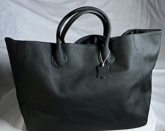 May include: A large, black leather tote bag with two rounded handles and a textured surface. The bag has a spacious interior and a small leather tag attached to the handle. The bag is designed for carrying items.