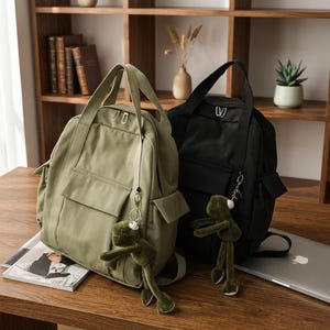 May include: Two backpacks, one sage green and one black, each featuring a top handle, a front pocket, and a plush frog keychain attached to the zip. The backpacks have a structured design with side pockets and silver-toned metal zip pulls.