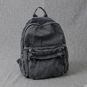 Può includere: Uno zaino in denim grigio antracite caratterizzato da un ampio scomparto principale con chiusura a zip, una tasca frontale più piccola con zip orizzontale, una maniglia superiore e dettagli con cuciture visibili su tutta la struttura.