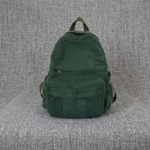 May include: A dark green canvas backpack featuring a large main compartment, a smaller front zipped pocket, and side pockets. The bag includes a top carry handle and adjustable shoulder straps, constructed from a durable, textured fabric material.