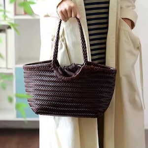 Pode incluir: Uma bolsa tote de couro trançado marrom escuro com alças trançadas. A bolsa tem formato retangular com fecho de cordão. A bolsa é segurada por uma pessoa que usa um casaco de cor clara.