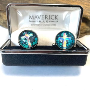 May include: A pair of cufflinks with a cross design, presented in a black and silver box. The cufflinks feature a teal background with a cross design. The box is labeled "MAVERICK MAN" and includes a website address.
