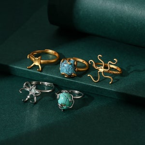 May include: Five gold and silver rings with turquoise stones and intricate designs. The rings feature a variety of shapes, including a cat, an octopus, and a flower.