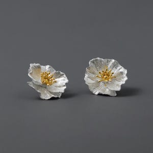 Puede incluir: Un par de pendientes de plata con forma de flor y centros dorados. Las flores tienen un aspecto texturizado y ligeramente arrugado.