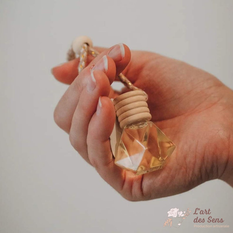 May include: A clear glass bottle with a geometric shape, filled with a light yellow liquid. The bottle is attached to a wooden ring and a string, making it a car air freshener. The bottle is held in a hand.