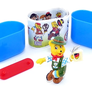 Könnte beinhalten: Eine gelbe Bärenfigur, die einen grünen Hut, ein blau-weißes Hemd und grüne Lederhosen trägt. Der Bär hält eine weiße Tasse mit einer deutschen Flagge. Die Figur ist Teil eines Sets von Sammelfiguren.
