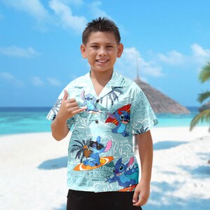 Op de afbeelding: Een lichtblauw overhemd met korte mouwen met een tropische print met Stitch van Lilo & Stitch. De print toont Stitch surfen op golven met palmbomen op de achtergrond.