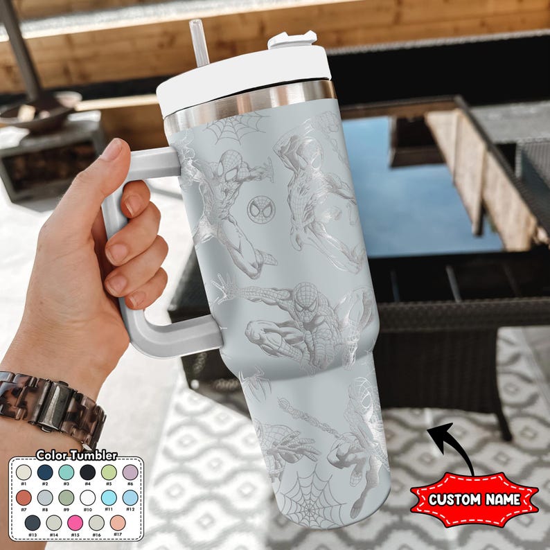 May include: A light gray tumbler with a white handle, lid, and straw. The tumbler features multiple illustrations of Spider-Man and a Spider-Man logo. The bottom right corner has a red banner that says "CUSTOM NAME".