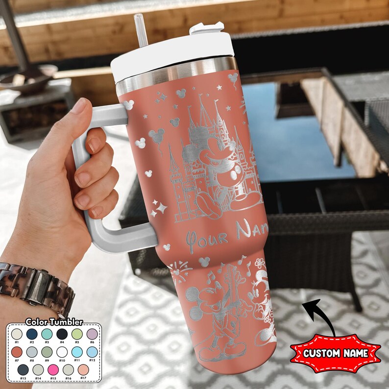 May include: A coral-colored tumbler with a white handle, lid, and straw. The tumbler features a silver engraved design of Mickey Mouse, a castle, and the words "Your Name". A color chart is visible.
