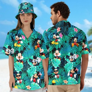May include: A matching set of a short-sleeved button-down shirt and a bucket hat. The teal fabric is covered in a pattern of Mickey Mouse, tropical leaves, and pink flowers. The shirt is worn by a person.