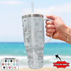 May include: A light gray tumbler with a white handle and lid, featuring engraved images of Minnie Mouse, Mickey Mouse, and a castle. The tumbler includes a straw and the text "YOUR NAME".