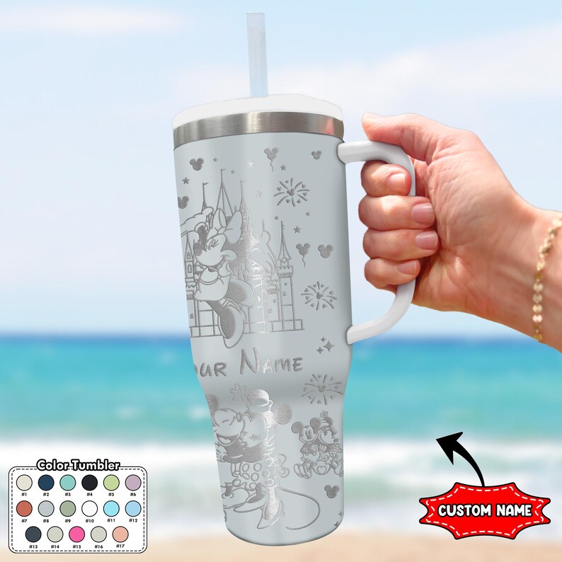 May include: A light gray tumbler with a white handle and lid, featuring engraved images of Minnie Mouse, Mickey Mouse, and a castle. The tumbler includes a straw and the text "YOUR NAME".