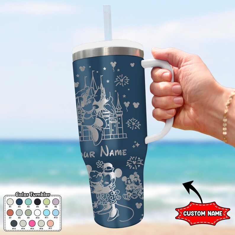 May include: A navy blue tumbler with a white handle and straw. The tumbler features a silver engraving of a castle, fireworks, and cartoon characters. The text "YOUR NAME" is visible. The tumbler is held by a person on a beach.