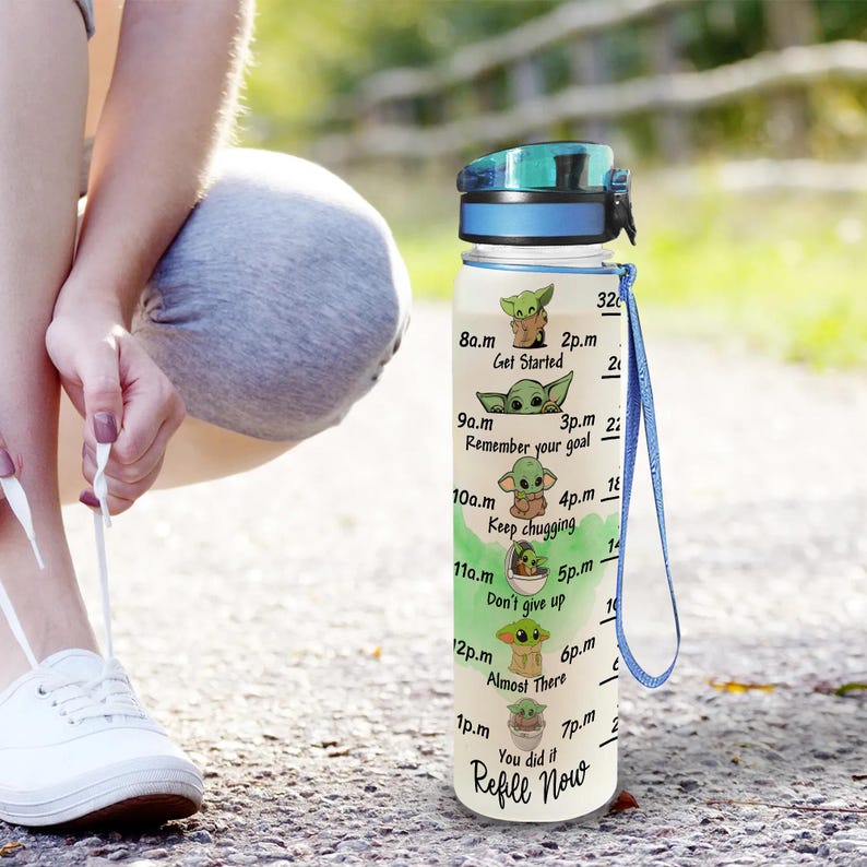 May include: A white water bottle with a blue lid and strap. The bottle has a motivational quote and images of a green alien character on it. The quote says "Refill Now" and includes times from 8 a.m. to 7 p.m. with a different image of the alien character for each time.