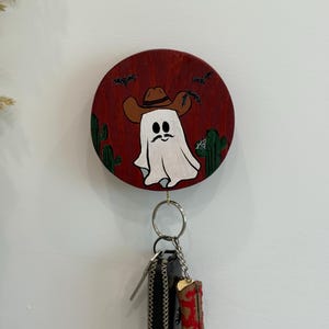 May include: A round, wooden key holder with a painted design of a ghost wearing a cowboy hat, set against a red background with cacti and bats. A key ring holds a black key fob and a miniature cowboy boot.