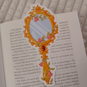 May include: A decorative bookmark shaped like a gold-colored mirror with a handle. The mirror has a floral design with pink flowers and green leaves. The bookmark is placed on top of an open book.
