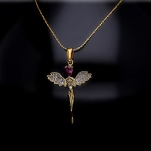 Puede incluir: Un collar de oro con un colgante que representa una figura de ángel alado con una piedra preciosa roja en el centro. El ángel está adornado con pequeñas piedras transparentes.